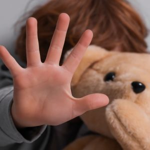 Mano abierta de un niño en primer plano en señal de “alto” o “no”, con un peluche desenfocado detrás, representando la protección y el rechazo al abuso infantil.